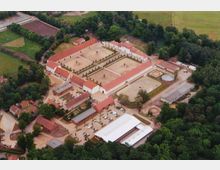 Luftaufnahme eines großen rechteckigen Gutshofs mit roten Dächern, umgeben von Wäldern und Wiesen. Der Innenhof verfügt über symmetrisch angeordnete Bäume und Brunnen, während umliegende Gebäude landwirtschaftlich genutzt werden.