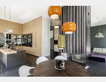 Modernes Wohn- und Esszimmer mit offener Küche, Holzelementen und schwarzen Oberflächen. Im Vordergrund ein runder Holztisch mit einer Affenskulptur, dahinter eine Küchenzeile mit Holzverkleidung sowie eine moderne Hängeleuchte mit farbigen Lampenschirmen und ein Kamin mit gestapeltem Brennholz.