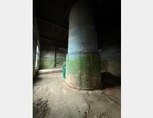 Innenraum eines verlassenen Industriegebäudes mit großem zylindrischem gemauertem Schornstein oder Ofen in der Mitte, unten grün gestrichen und oben grau. Umgeben von rauen Backsteinwänden, Betonboden mit Schmutz und Rissen sowie hohen Fenstern an der linken Seite unter einem Holzdach.