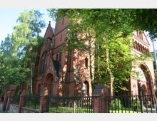 Historische Backsteinkirche mit spitzbogigen Eingangsportalen und verzierten Fenstern, umgeben von Bäumen und einem schmiedeeisernen Zaun. Die Fassade zeigt rote Ziegelarchitektur mit gotischen Elementen und kleineren Statuen.
