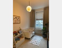 Kinderzimmer mit hellem Teppichboden, einem weißen Babybett, einem Bücherregal mit Spielzeug und Büchern, einem Alphabet-Teppich auf dem Boden und einem großen Fenster mit beigen Vorhängen. Eine runde Hängelampe und eine Zimmerpflanze ergänzen die Einrichtung.