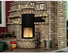 Ein gemauerter Kamin aus hellen Backsteinen mit einem schwarzen Kaminofen, in dem ein Feuer brennt. Um den Kamin stehen Zubehör wie ein Metalleimer, Kaminbesteck und Holzscheite, daneben ein roter Eimer und weitere Werkzeuge.