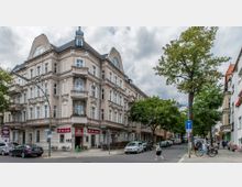 Eckgebäude im historischen Baustil mit hellem Putz und dekorativen Verzierungen, darunter Balkone und ein gewölbtes Dachfenster; im Erdgeschoss befinden sich Geschäfte und ein Café mit Außenbestuhlung. Eine straßenbegleitende Baumreihe sowie geparkte Autos sind auf beiden Straßenseiten zu sehen.