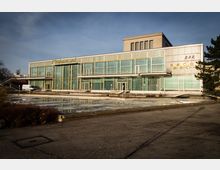 Ein modernistisches Gebäude mit großen Glasfronten und vertikalen Rahmen, beschriftet mit "Restaurant" und "Bar" an der Fassade. Davor befindet sich ein leerer, gepflasterter Platz mit einem dekorativen Wasserbecken.