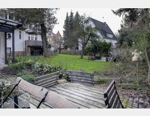 Ein grüner Garten mit Rasenfläche, umgeben von Bäumen und Sträuchern, im Hintergrund Wohnhäuser mit Satteldächern. Im Vordergrund sind ein Holztisch und Bänke auf einem gepflasterten Bereich zu sehen.