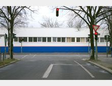Eine Kreuzung mit Verkehrsampeln, umgeben von kahlen Bäumen, vor einem flachen Gebäude mit weißer Fassade und blauer Basis, das durch eine rote Linie abgesetzt ist. Im Vordergrund verlaufen Fahrbahnmarkierungen.