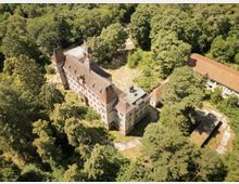 Luftaufnahme eines historischen Schlosses mit rotem Ziegeldach und zwei markanten Türmen, umgeben von dichtem Wald. Im Hintergrund befinden sich ein weiteres Gebäude mit rotem Dach und eine freie Fläche mit Wegen.