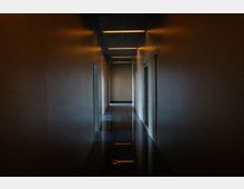 Langer, schmaler Innenflur in einem modernen Gebäude mit glatten Wänden, mehreren Türen an beiden Seiten und einem dunklen, glänzenden Fliesenboden. An der abgehängten Decke verlaufen rechteckige Leuchtstreifen, die sich im Boden spiegeln.