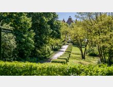 Ein Weg, gesäumt von niedrigen Hecken, führt durch eine parkähnliche Landschaft mit hohen Bäumen; im Hintergrund ist ein historisches Gebäude mit roten Dächern und markanten Türmen sichtbar. Links im Bild steht ein Schild mit der Aufschrift „Bismarckturm“.