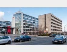 Straßenansicht mit Bürogebäuden; links ein modernes weißes Gebäude mit Glasfassade und roten Akzenten, daneben ein schlichtes graues Gebäude mit rechteckigen Fenstern. Im Vordergrund eine befahrene Kreuzung mit geparkten und fahrenden Autos.