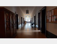 Langer, schmaler Flur in einem historischen Gebäude mit Holztüren auf beiden Seiten, glänzendem Boden und hölzernem Mobiliar; am Ende des Flurs befindet sich ein großes Fenster mit dekorativen Glasdetails und Sitzmöbeln.