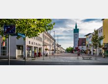 Städtischer Platz mit Rathausgebäude links und Uhrturm mit grünem Dach im Hintergrund. Der Bereich ist von modernen Gebäuden, Schildern, Straßenlaternen, Bäumen und Blumenampeln gesäumt.