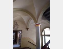 Treppenhaus in einem historischen Gebäude mit Gewölbedecken, einer verzierten Säule und verblassten Wandmalereien. Rechts befindet sich ein großes Fenster mit Bögen, links ein offener Durchgang mit freiliegendem Mauerwerk.