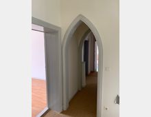 Fluransicht in einem Innenraum mit Spitzbogen-Durchgang aus grauem Holzrahmen; rechts ein Lichtschalter an einer cremefarbenen Wand, der Boden ist mit beigefarbenem Teppich ausgelegt, daneben ein Raum mit laminierter Holzoptik.