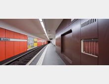 U-Bahn-Station mit einem Bahnsteig, der von orangefarbenen Wandpaneelen und Werbeplakaten gesäumt ist; auf der rechten Seite befinden sich braune Wandverkleidungen mit eingelassenen Fenstern. Eine gebogene Schiene verläuft in die Ferne, unter einer hellen Deckenbeleuchtung.