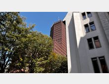 Eine Außenaufnahme mit einem modernen roten Hochhaus im Hintergrund, teils verdeckt durch grüne Bäume. Im Vordergrund ein weißes Gebäude mit großen Fenstern, beleuchtet von Sonnenlicht, vor klarem, blauem Himmel.