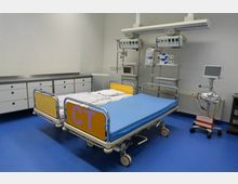 Krankenhauszimmer mit einem Bett auf Rollen, blauer Matratze und gelben Seitenelementen, darauf die Aufschrift „CT“. An der Wand befinden sich medizinische Geräte, Monitore und Hängeschränke. Der Boden ist blau, die Wände sind weiß.