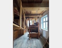 Holzinterieur einer historischen Mühle mit sichtbarer Holzkonstruktion, alten Holzböden und einer traditionellen Mühlenmaschine. Tageslicht fällt durch zwei Fenster mit weißem Rahmen an der rechten Wand.