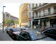 Straßenszene in einer Stadt mit parkenden Autos am Straßenrand, gelben und cremefarbenen Wohnhäusern mit dekorativen Fassaden sowie Balkonen mit Pflanzen. Im Hintergrund ist ein historisches Gebäude mit großen Fenstern und rotem Dach erkennbar, begleitet von Baustellenaktivitäten und Bäumen.