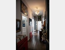 Ein schmaler, geschmackvoll dekorierter Flur mit Holzboden und weiß gestrichenen Wänden. An den Wänden hängen Bilder, und Möbelstücke, wie eine rote Kommode und ein Holztisch mit Dekorationen, stehen entlang des Ganges; eine kugelförmige Deckenleuchte hängt von der Decke.