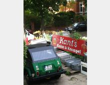 Ein kleiner Hinterhof mit einem alten, grünen Fahrzeug mit schwarzem Verdeck im Vordergrund. Im Hintergrund ein rotes Schild mit der Aufschrift „Kant's Kunst & Krempel“, umgeben von verschiedenen Objekten wie Metallwannen und Holzpaletten; im Hintergrund stehen Bäume und ein Backsteingebäude.