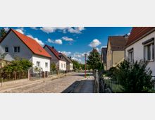 Straße in einem Wohngebiet mit freistehenden Einfamilienhäusern, die rote oder dunkle Schrägdächer haben. Die gepflasterte Straße wird von Gehwegen und Vorgärten mit Bäumen und Büschen gesäumt; der Himmel ist blau mit vereinzelten Wolken.