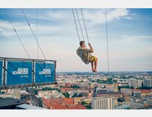 Ein Mann schaukelt an einer hohen Schaukelplattform mit Aufschrift „Jochen Schweizer Base Flying“ über einer Großstadt, mit Blick auf Dächer, Kirchen und moderne Gebäude unter einem leicht bewölkten Himmel.