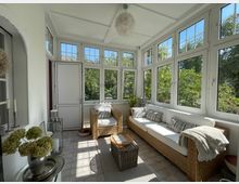 Wintergarten mit großen, weißen Sprossenfenstern, die viel Tageslicht hereinlassen. Der Raum ist mit Rattanmöbeln, weißen Polstern und dekorativen Kissen eingerichtet, mit Blick auf einen grünen Garten.