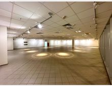 Großer, leerer Innenraum mit gefliesten Böden und einer Rasterdecke mit eingebauten Leuchten und Belüftungselementen. Die Wände sind weiß, und vereinzelte Lichtkreise sind auf dem Boden erkennbar.