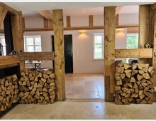 Innenraum eines rustikalen Gebäudes mit sichtbaren Holzbalken und gestapeltem Kaminholz auf beiden Seiten eines offenen Durchgangs. Der Boden ist mit beigen Steinfliesen ausgelegt, und durch Fenster fällt Tageslicht in den Raum.