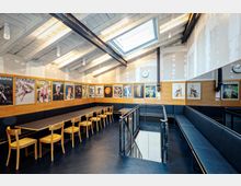 Ein heller, modern gestalteter Aufenthaltsraum mit weiß getäfelter Holzdecke, Oberlichtfenster und einer langen Tischreihe mit Holzstühlen. An den Wänden befinden sich Holzvertäfelungen mit Postern, und ein Treppenabgang mit schwarzem Metallgeländer ist im Vordergrund sichtbar.