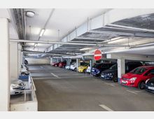 Ein mehrstöckiges Parkhaus mit parkenden Autos in einer offenen Parkreihe. Die Decke zeigt sichtbare Rohre und Beleuchtung, ein Einfahrt-verboten-Schild ist an einer Fahrbahnmarkierung angebracht.