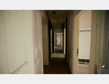 Ein schmaler, leerer Flur in einem verfallenen Gebäude mit abblätternden Wänden und mehreren offenen Türen. Im Hintergrund sind weitere Räume und ein Bereich mit weiß gefliesten Wänden sichtbar.