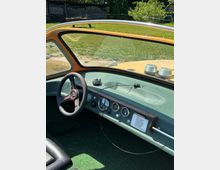 Blick aus dem Innenraum eines Oldtimers auf Armaturenbrett und Lenkrad, mit analogen Rundinstrumenten und einem kleinen Handschuhfach. Durch die gewölbte Frontscheibe sind die Motorhaube sowie ein grüner Garten im Hintergrund zu sehen.