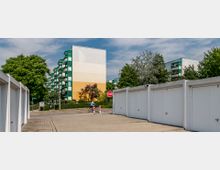 Mehrstöckige Wohngebäude mit bunten Fassaden und grünem Balkonrand, umgeben von Bäumen und Sträuchern. Im Vordergrund befinden sich weiße Garagen und ein gepflasterter Weg, auf dem eine Person mit Fahrrad unterwegs ist, sowie ein Stoppschild.