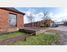 Industriegelände mit historischen Backsteingebäuden. Im Vordergrund eine niedrige Plattform aus Backstein, daneben Rasen und ein alter Rohrbogen; im Hintergrund weitere Backsteinbauten mit großen Fenstern sowie ein gelbes Fahrzeug und Baugeräte.