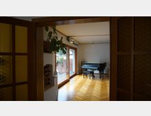 Blick in ein Wohnzimmer mit Holzboden und großen Fensterfronten, die zu einer Terrasse mit Backsteinmauer führen. Im Raum steht ein schwarzer Flügel, daneben befinden sich Pflanzen und eine kleine Weinablage.