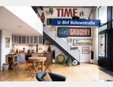 Offener Wohnbereich mit einer Küche in modernem Design, einem runden Esstisch aus Holz und dazu passenden Stühlen. Im Hintergrund ist eine Treppe zu sehen, deren Wand mit verschiedenen Retro-Schildern und Dekorationen gestaltet ist, darunter ein Schild mit der Aufschrift "U-Bhf Bülowstraße". Auf der rechten Seite stehen große Fenster, die viel Tageslicht hereinlassen.