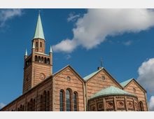 Backsteinkirche im neoromanischen Stil mit einer hohen Glockenturmspitze und grün patinierten Dächern. Die Fassade ist aus rotbraunem Ziegelstein gefertigt, mit Rundbogenfenstern und einem Uhrzifferblatt am Turm.