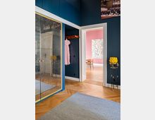 Innenraum einer Wohnung mit dunklen blauen Wänden und hellem Fischgrätenparkett. Zu sehen sind ein verspiegelter Einbauschrank mit goldenen Akzenten, ein offener Durchgang zu einem rosa gestrichenen Raum, und ein goldfarbener Beistelltisch mit gelben Dekoelementen.