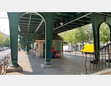 Straßenszene mit Überführung einer grünen stählernen Hochbahn und Pfeilern, darunter ein kleines rundes Gebäude mit Schaufenstern. Im Hintergrund geparkte Autos, Bäume und ein gelbes Lieferfahrzeug.