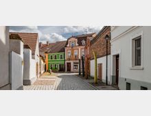 Gepflasterte Altstadtgasse mit niedrigen, farbenfrohen Häusern, darunter grüne und gelbe Fassaden. Rechts und links stehen Gebäude mit roten Ziegeldächern, und eine Straßenlaterne sowie Holzskulpturen säumen die Straße.