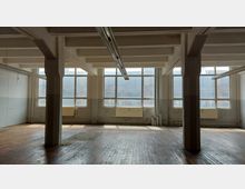 Innenansicht eines leeren, industriellen Raums mit Holzboden, großen Fenstern mit Metallrahmen und sichtbaren weißen Stahlträgern an der Decke. Heizkörper und Rohre sind an den Wänden montiert, und die Fenster lassen gedämpftes Tageslicht herein.