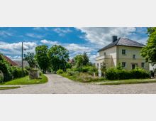 Straßenszene in einer ländlichen Wohngegend mit gepflastertem Weg, einem gelben Einfamilienhaus mit Walmdach rechts und dichter Vegetation links und im Hintergrund. Einige Bäume und ein Strommast sind ebenfalls zu sehen, sowie ein kleiner Kasten mit Graffiti.