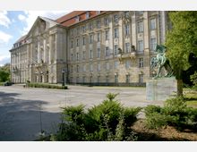Neoklassizistisches Gebäude mit detaillierter Fassade, hohen Fenstern und rotem Mansarddach. Rechts im Vordergrund steht eine Reiterstatue auf einem Podest, umgeben von Grünflächen und Bäumen.