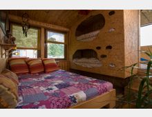 Schlafzimmer in einer Holzhütte mit Wänden und Decke aus hellem Holz. Zu sehen sind ein großes Bett mit bunt gemusterter Bettdecke und Kissen sowie eingebaute Etagenbetten aus Spanplatte mit organisch geformten Ausschnitten. Durch die Fenster fällt Tageslicht und es gibt einen Ausblick auf grüne Bäume.
