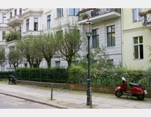 Mehrstöckige Wohngebäude mit hellen Fassaden im Altbaustil; davor eine Reihen von Bäumen, ein schmiedeeiserner Zaun und ein Gehweg mit Pflastersteinen. Am Straßenrand steht eine rote Vespa neben einer historischen Laterne.