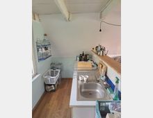 Kleine Küchenzeile in einem schmalen Raum mit Holzfußboden, weiß gestrichener Decke mit sichtbaren Balken und einer Hängelampe. Rechts sind Spüle, Abtropfgestell und Arbeitsfläche mit Küchenutensilien, links ein offenes Wandregal mit Gläsern sowie Wasserkisten am Boden unter einem Fenster.