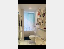 Kleines Badezimmer mit weißen Fliesen, einem Waschbecken, einer Toilette und einem Handtuchheizkörper. Über der Badewanne hängt ein blauer Duschvorhang, Regale an der Wand enthalten verschiedene Pflegeprodukte.