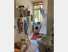 Ein kleines Badezimmer mit Fliesenboden, einer Dusche mit einem Vorhang im Fischdesign, einer Waschmaschine unter einem Fenster mit Blick ins Grüne und einem Waschbecken. An den Wänden befinden sich offene Regale mit Handtüchern und Pflegeprodukten sowie Haken mit hängenden Handtüchern.